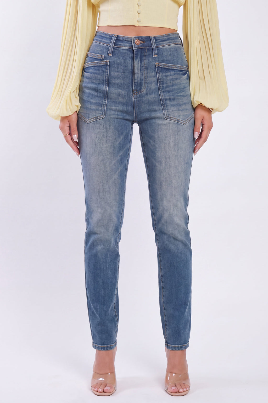 A model wearing a pair of high-waisted, straight-leg blue denim jeans with a faded wash, paired with a pale yellow long-sleeve top and clear high-heeled sandals, set against a plain white studio background.