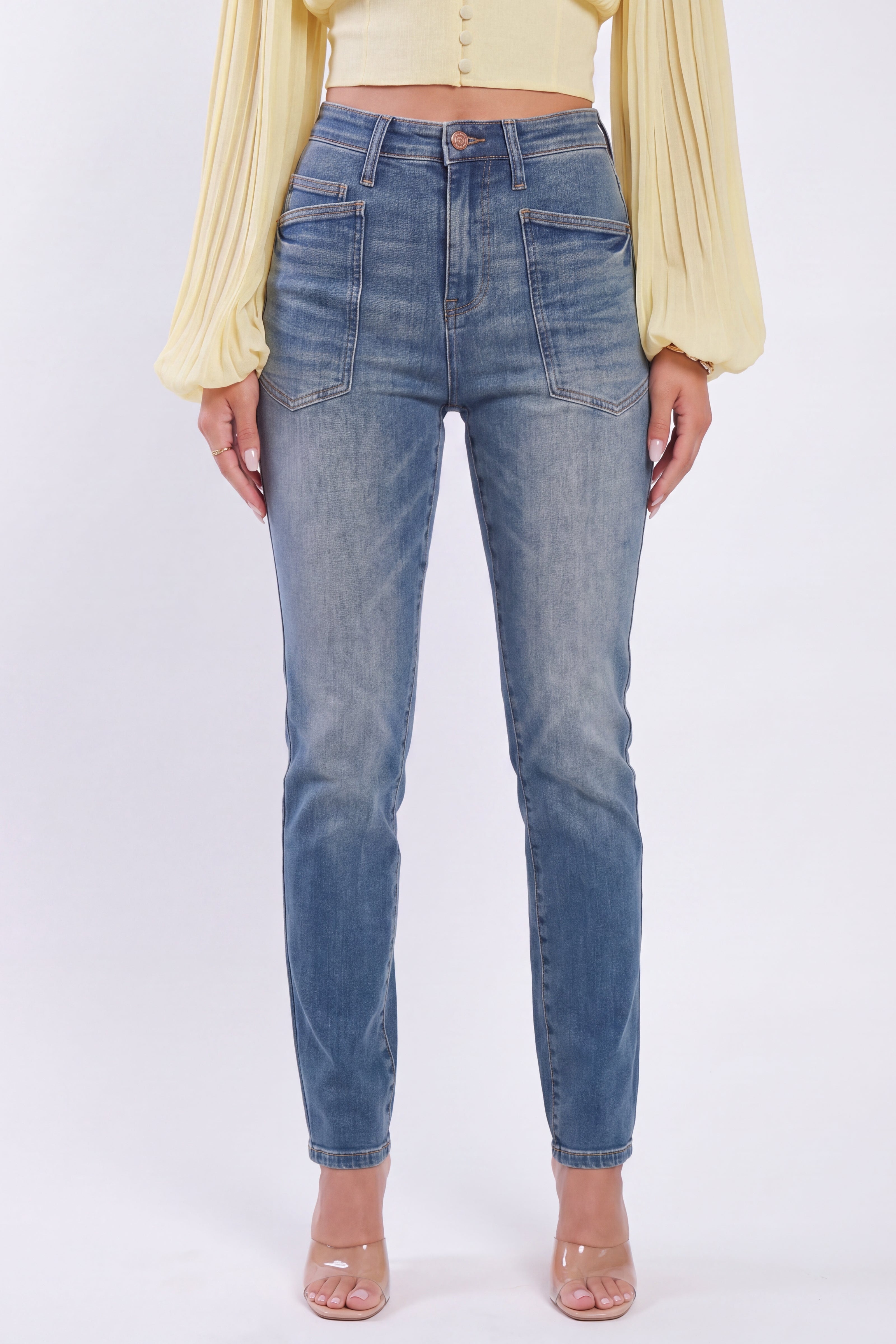 A model wearing a pair of high-waisted, straight-leg blue denim jeans with a faded wash, paired with a pale yellow long-sleeve top and clear high-heeled sandals, set against a plain white studio background.
