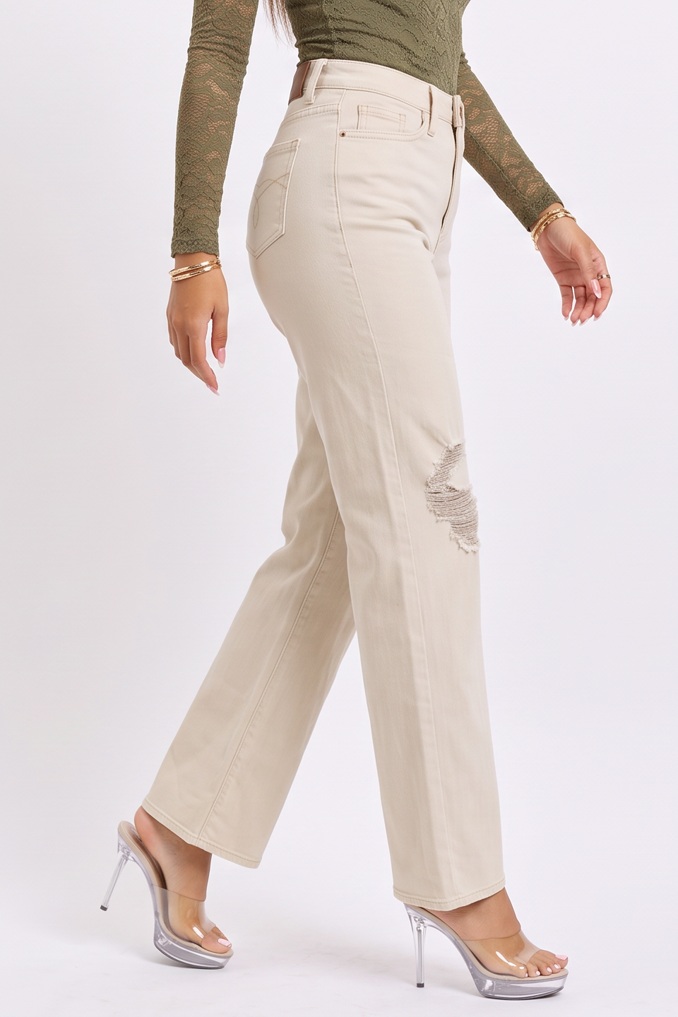 A model wearing a pair of off-white, high-waisted straight-leg jeans with distressed details on the knees, styled with a green long-sleeve top and clear high-heeled sandals against a plain white studio background.