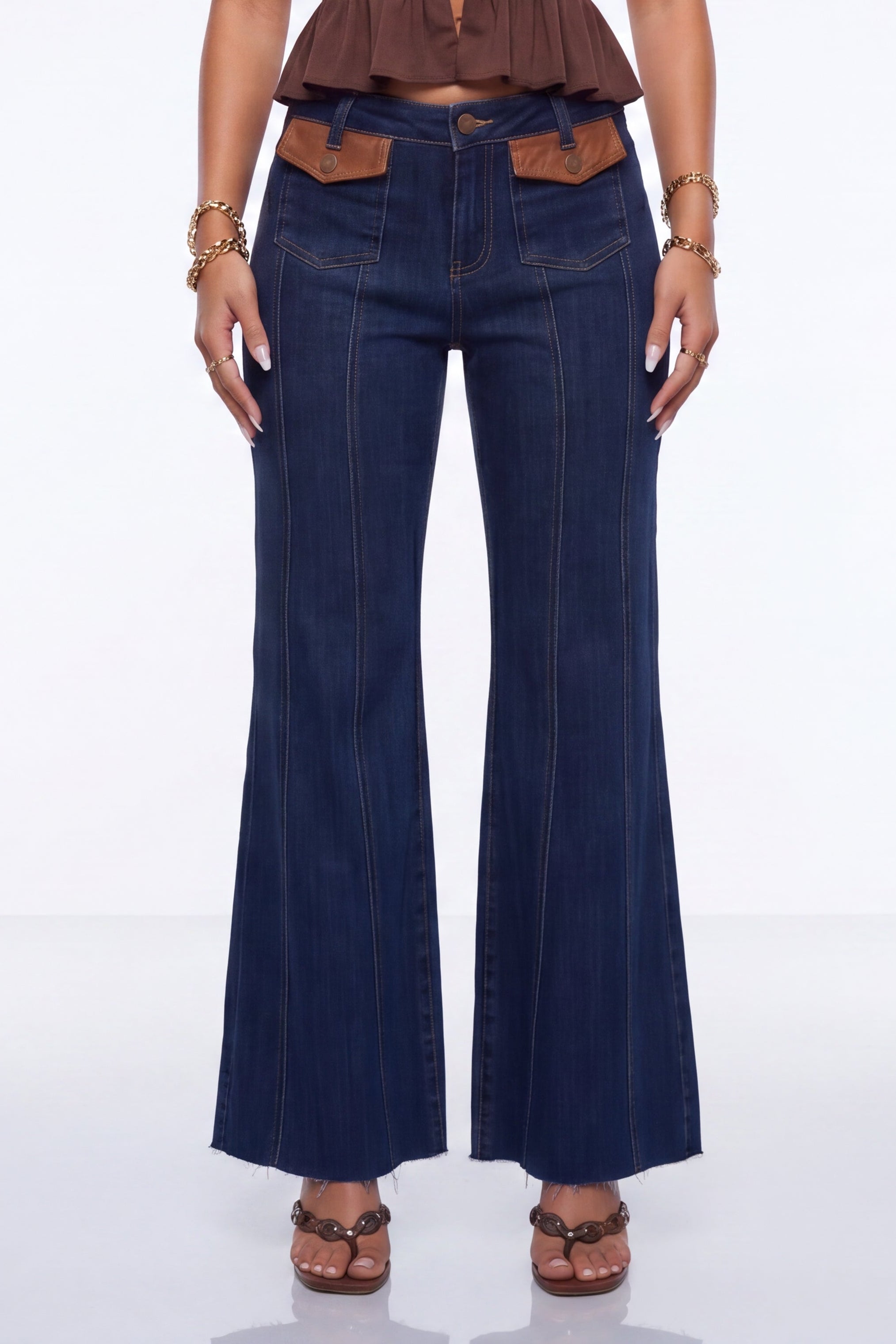 A model wearing dark blue flare jeans with brown leather accents at the waistband and brown sandals, shown from the waist down against a white studio background.