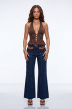 A model stands against a white studio background wearing a brown halter-neck wrap top with a deep V-neckline and gathered waist, paired with dark blue wide-leg jeans featuring brown leather-like accents at the waistband, gold jewelry, and brown sandals.