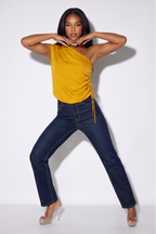 A woman modeling a mustard yellow one-shoulder ruched top and dark blue jeans against a plain white studio background.