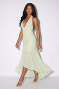 A woman modeling a pale mint green halter-neck midi dress with a deep V-neckline and a flowing asymmetrical hem, set against a plain white studio background.