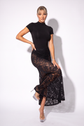 A woman modeling a black short-sleeve mock-neck bodysuit with a sheer lace overlay skirt, paired with clear high-heeled sandals, against a white studio background.