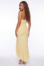 A woman modeling a pale yellow strapless maxi dress with a ruched bodice, back cutout, and flared ruffled hem, shown against a plain white studio background.